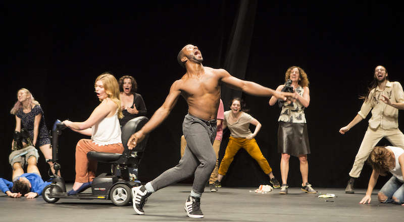 A cast of characters of different shapes and sizes smile and dance onstage. A shirtless man opens his mouth wide is at the center.